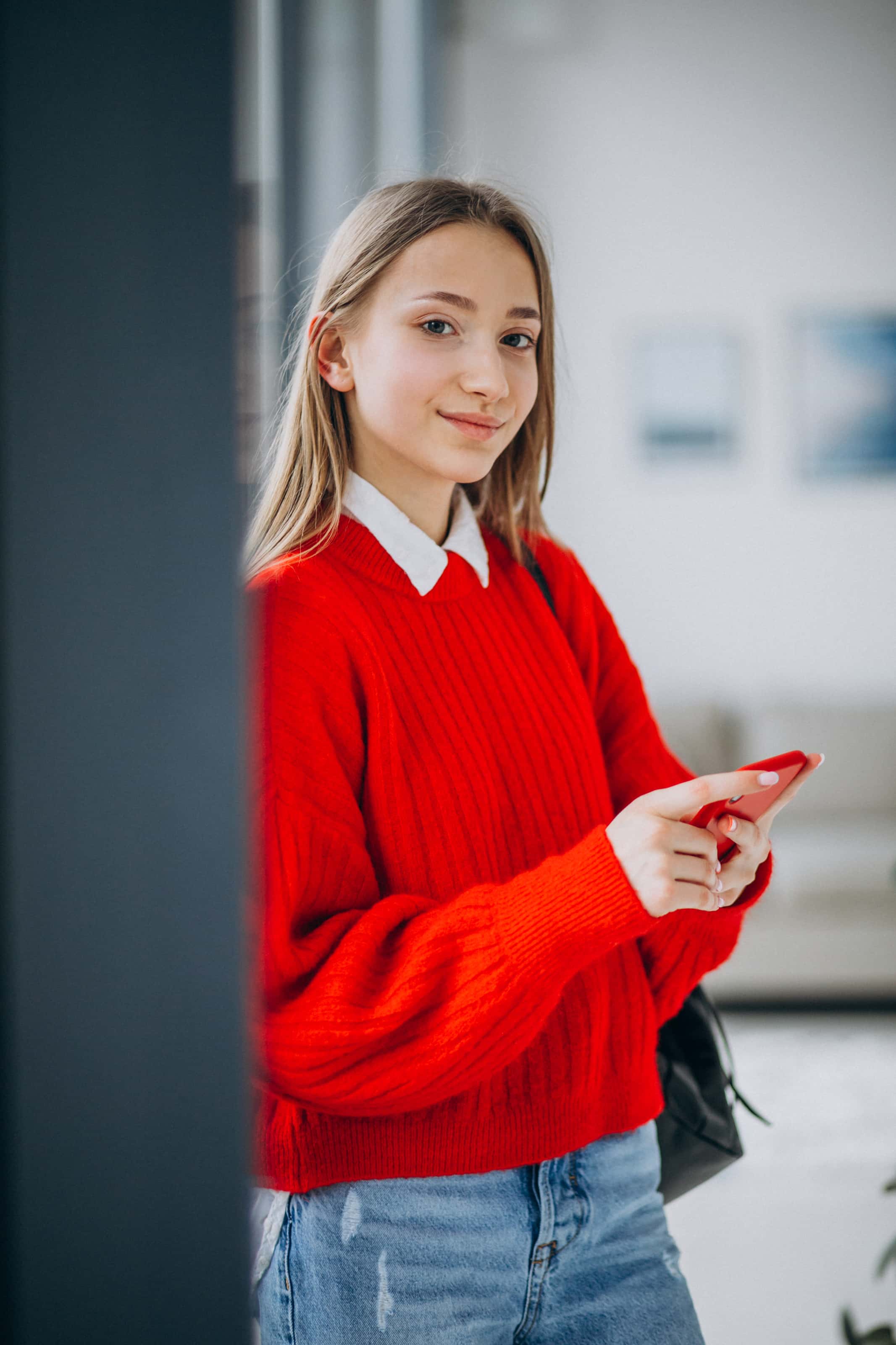 Nastolatka w czerwonym swetrze trzymająca smartfon, uczennica o profilu humanistycznym zainteresowana językiem i kulturą.