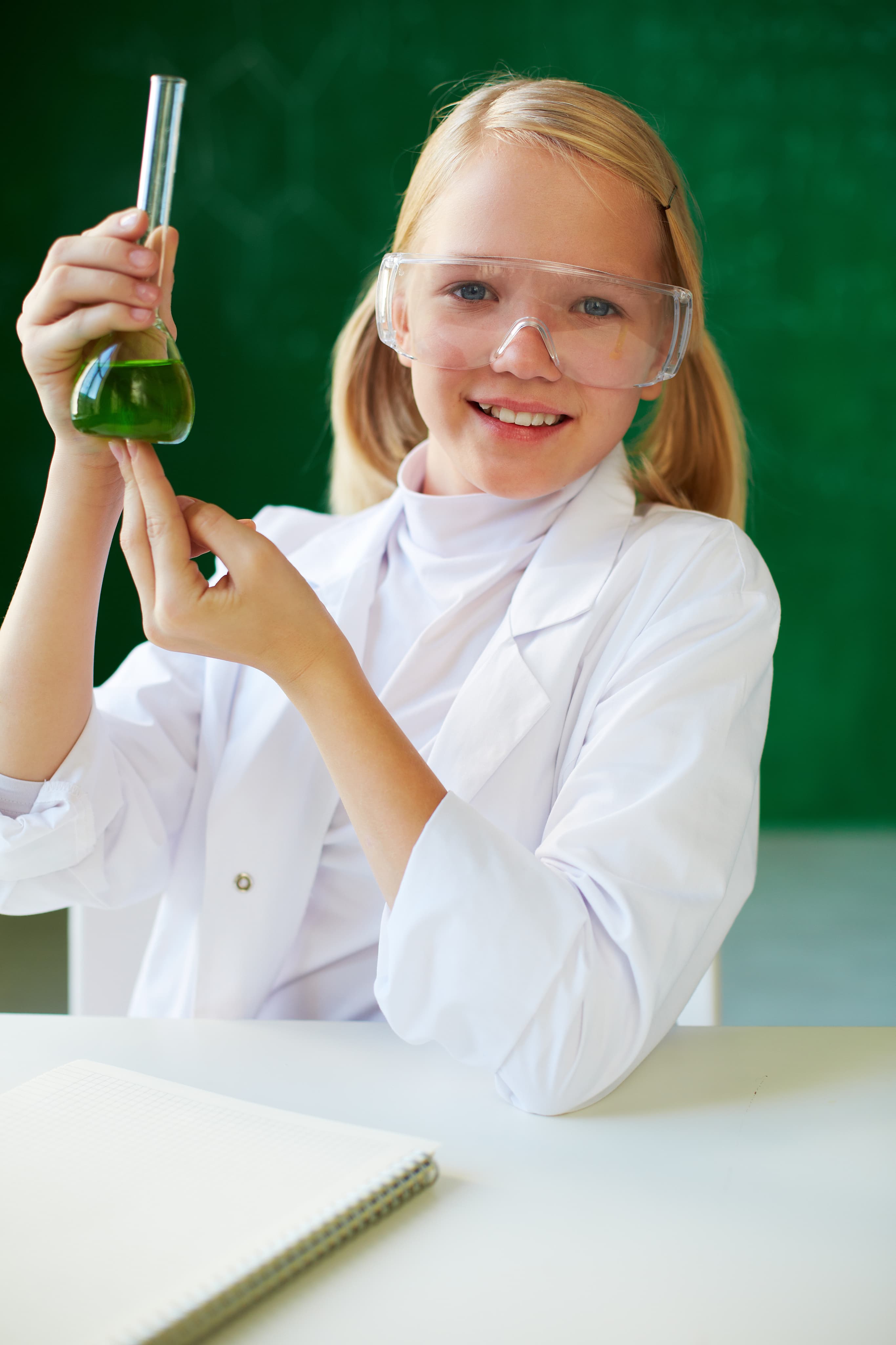 Dziewczynka w fartuchu laboratoryjnym trzyma zielony roztwór w kolbie, eksperyment chemiczny na lekcji biologii i chemii.